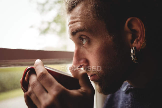 Nahaufnahme eines jungen Mannes, der zu Hause durch ein Fenster Kaffee trinkt — Stockfoto