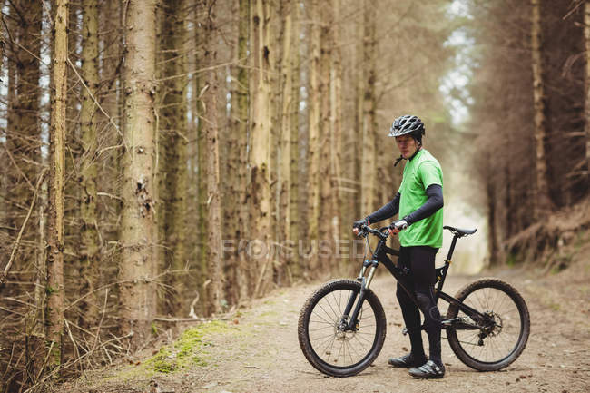 Vista lateral del ciclista de montaña de pie junto a los árboles en el bosque - foto de stock