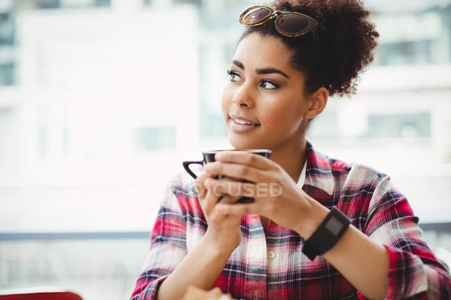Mujer sonriente reflexiva sosteniendo la taza de café mientras está sentado en la mesa en el restaurante - foto de stock