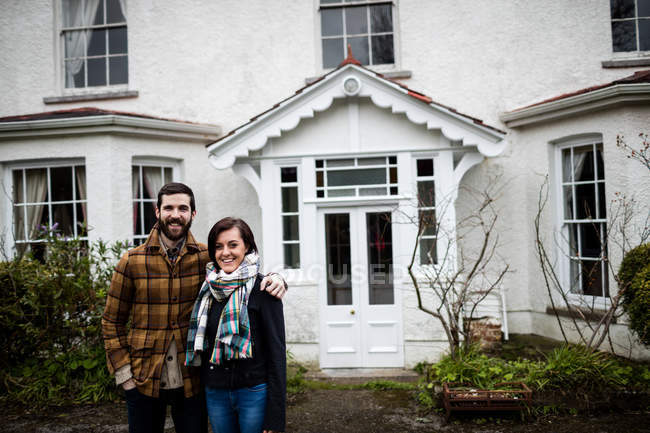 Portrait de couple debout près d'une nouvelle maison avec les bras autour — Photo de stock