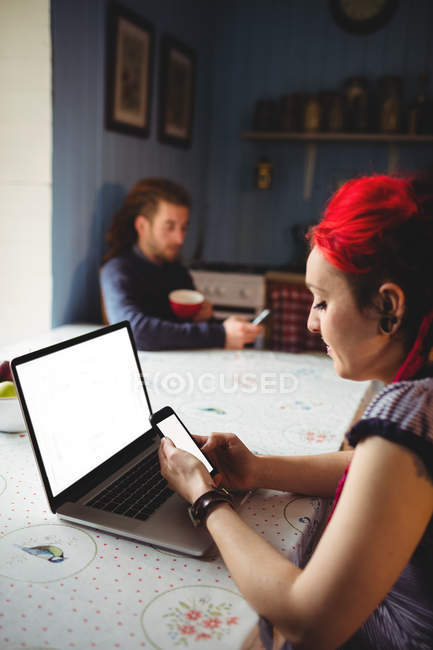 Junge Frau benutzt Handy und Laptop, während Mann im Hintergrund zu Hause ist — Stockfoto