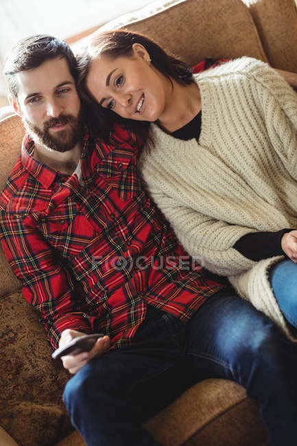 Couple sitting on sofa with remote control while watching television in living room at home — Stock Photo