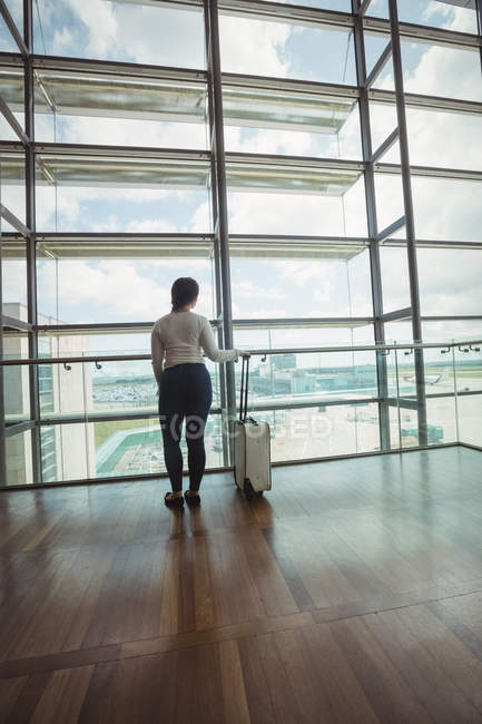 Rückansicht einer Geschäftsfrau mit Gepäck, die durch ein Glasfenster am Flughafen blickt — Stockfoto