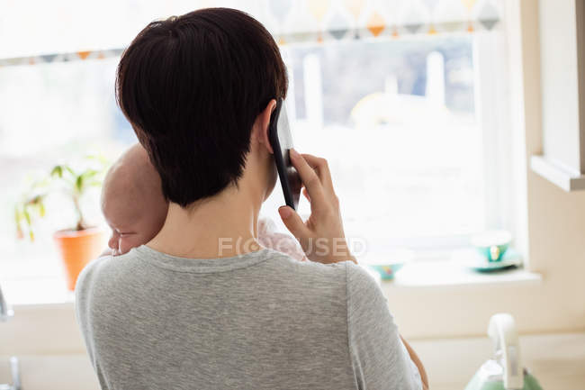 Visão traseira da mãe segurando seu bebê enquanto conversa por smartphone na cozinha em casa — Fotografia de Stock