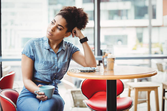 Mujer inteligente reflexiva sosteniendo la taza de café mientras está sentado en la mesa en el restaurante - foto de stock