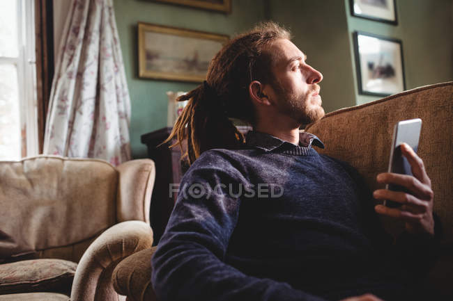 Jeune homme hipster tenant un téléphone portable sur le canapé à la maison — Photo de stock