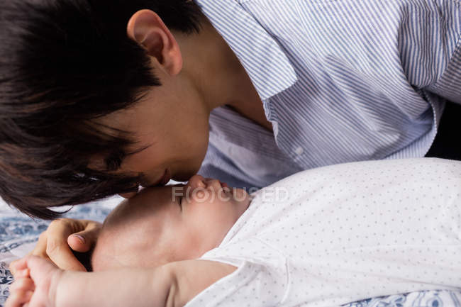 Madre besando su frente de bebé mientras él duerme en la sala de estar en casa - foto de stock