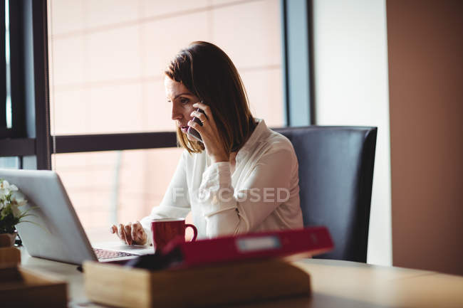 Mujer de negocios usando el ordenador portátil y hablando en el teléfono móvil en la oficina - foto de stock