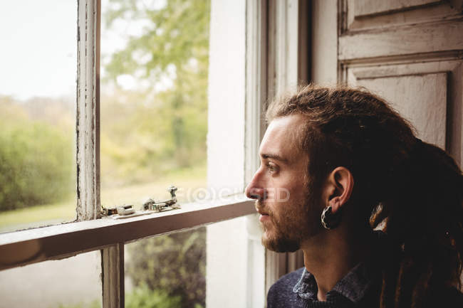 Vista lateral del hipster mirando a través de la ventana en casa - foto de stock