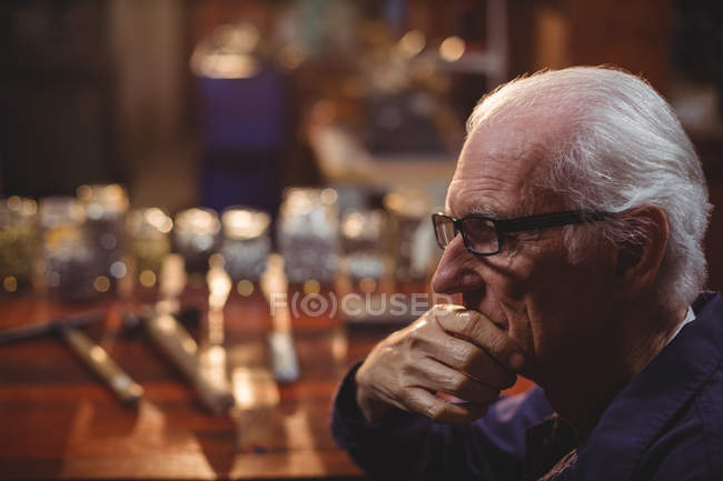 Thoughtful shoemaker sitting in workshop with hand on chin — Stock Photo