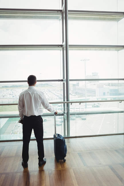 Вид ззаду бізнесмена з багажем, який дивиться через скло в аеропорту — стокове фото