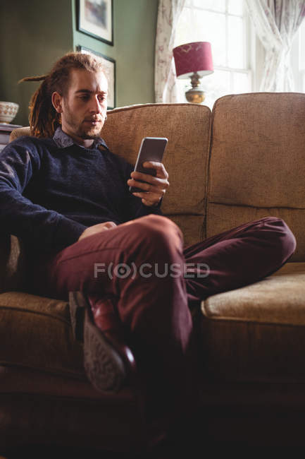 Jeune homme utilisant un téléphone portable assis sur le canapé à la maison — Photo de stock