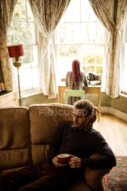 Vista de ángulo alto del hombre con taza de café en el sofá, mientras que la mujer en el fondo en casa - foto de stock