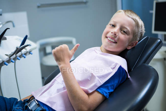 Sonriendo joven paciente sentado en la silla del dentista y mostrando sus pulgares hacia arriba en la clínica - foto de stock