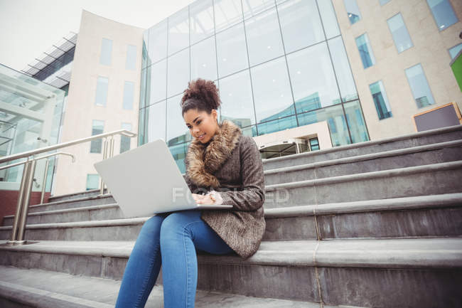 Низкий угол обзора женщины с ноутбуком, сидящей на ступеньках против современного здания — стоковое фото