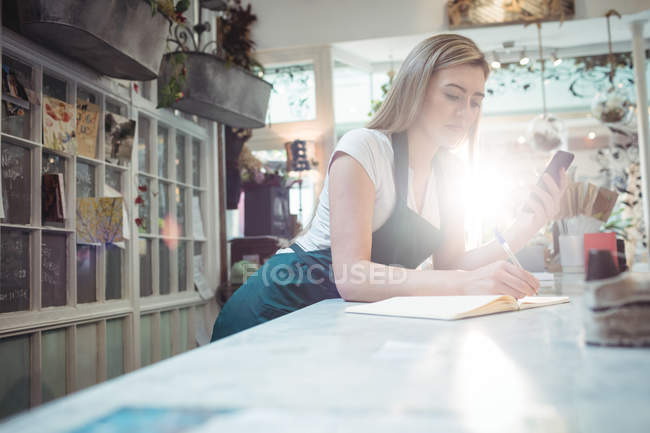 Florista feminina observando no diário enquanto segurava o telefone celular na loja de flores — Fotografia de Stock