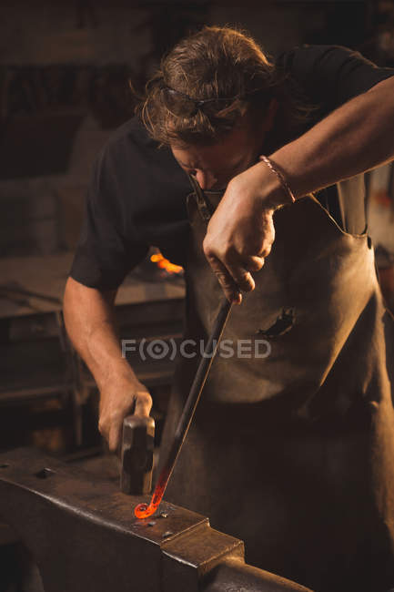 Herrero trabajando en metal caliente usando martillo para dar forma en el taller - foto de stock