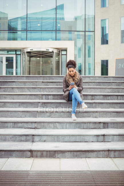 Повна довжина молодої жінки тримає телефон, сидячи на сходах — стокове фото