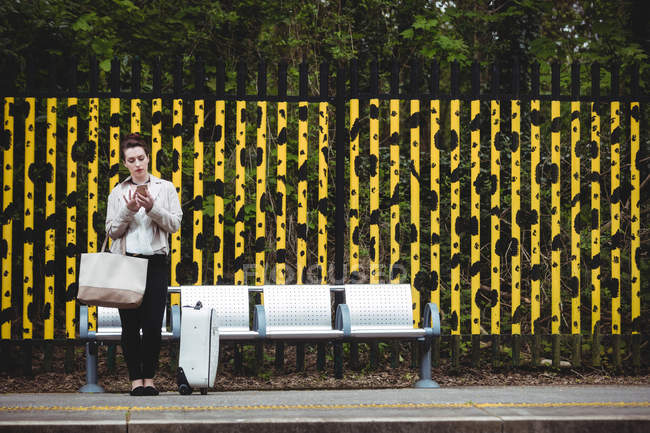 Молодая женщина использует мобильный телефон против забора на железнодорожной станции платформы — стоковое фото