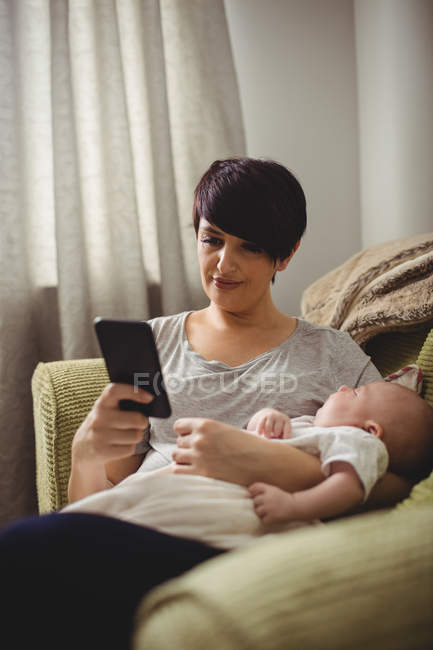 Mère utilisant un smartphone pendant que le bébé dort dans son bras au salon — Photo de stock