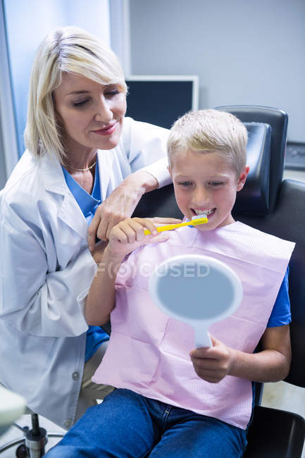 Dentista asistiendo a paciente joven mientras se cepilla los dientes en la clínica dental - foto de stock
