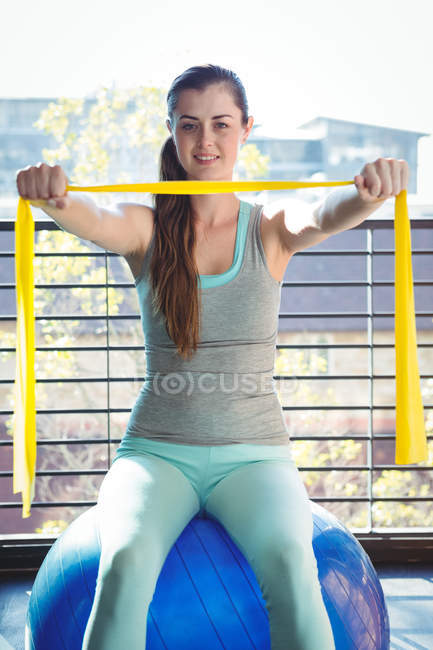 Retrato de la mujer sosteniendo la banda de resistencia mientras está sentado en la pelota de ejercicio en el gimnasio - foto de stock
