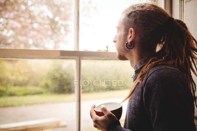 Seitenansicht des Hipsters, der eine Kaffeetasse hält, während er zu Hause durch das Fenster schaut — Stockfoto