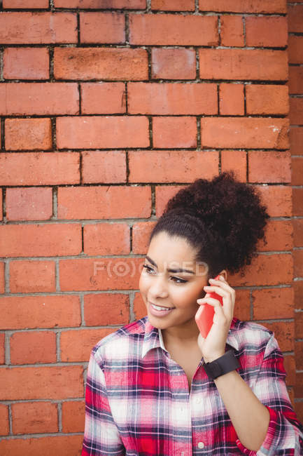 Mujer bonita sonriente hablando por teléfono mientras está de pie contra la pared de ladrillo - foto de stock