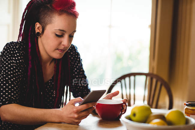 Jovem mulher usando telefone enquanto se senta à mesa em casa — Fotografia de Stock