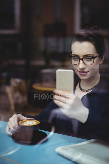 Young woman taking selfie in cafe seen through glass — Stock Photo