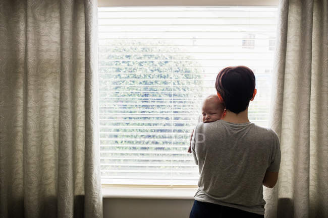Rückansicht der Mutter, die ihr kleines Baby hält und zu Hause durch das Fenster schaut — Stockfoto