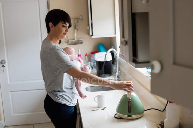 Мама держит своего малыша, пока делает кофе на электрическом чайнике на кухне — стоковое фото
