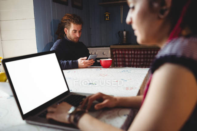 Молода жінка використовує ноутбук, а чоловік використовує мобільний телефон вдома — стокове фото