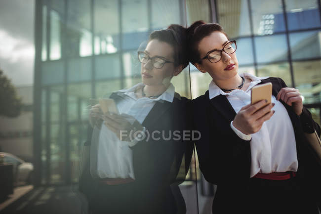 Молода бізнес-леді, використовуючи мобільний телефон, що відображає на склі, спираючись — стокове фото