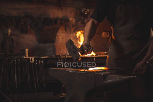 Herrero de sección media trabajando en metal caliente usando martillo para dar forma en el taller - foto de stock
