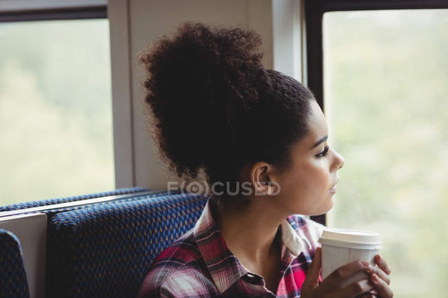 Крупный план вдумчивой женщины, смотрящей в окно, когда она пьет кофе в поезде — стоковое фото