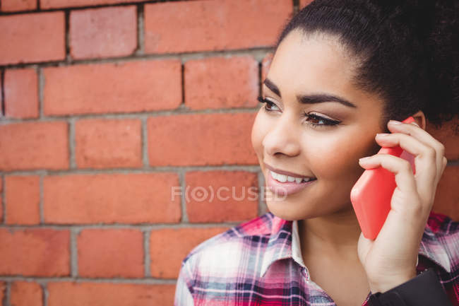 Close-up de mulher sorridente falando ao telefone enquanto está de pé contra a parede de tijolo — Fotografia de Stock
