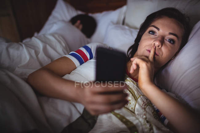 Ritratto di donna che usa il cellulare con un dito sulle labbra in camera da letto — Foto stock