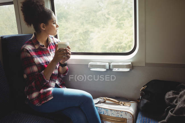 Jovem atenciosa olhando pela janela enquanto toma café no trem — Fotografia de Stock