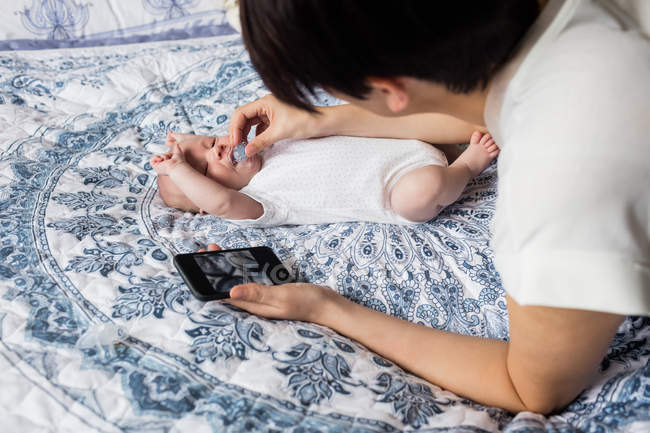Madre poniendo maniquí en su boca de bebé y sosteniendo el teléfono inteligente en casa - foto de stock