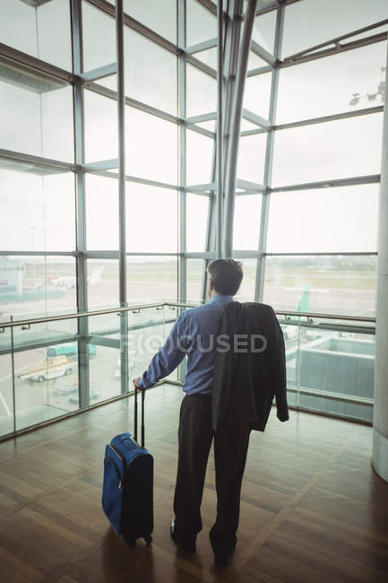 Вид сзади бизнесмена с багажом, смотрящего через стеклянное окно в аэропорту — стоковое фото