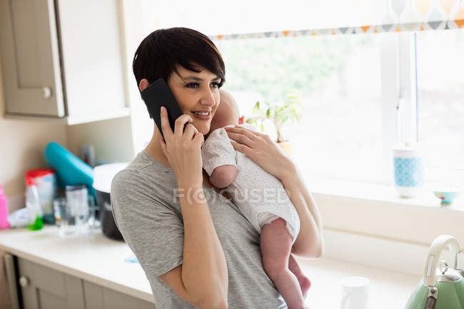 Мама держит своего малыша, разговаривая со смартфоном дома на кухне — стоковое фото