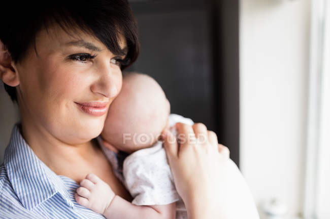 Madre de pie cerca de la ventana y sosteniendo a su bebé en casa - foto de stock