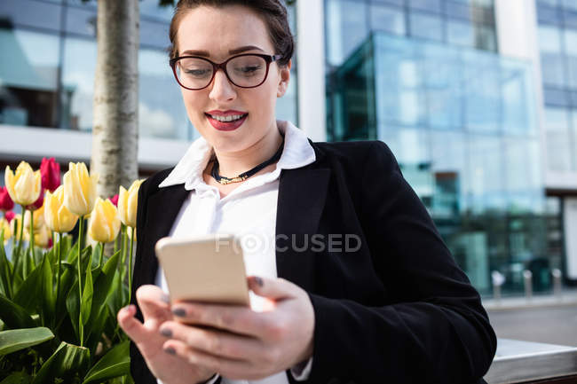 Smiling young businesswoman using mobile phone against office building — Stock Photo
