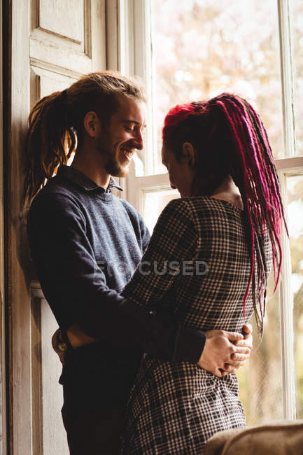 Feliz joven pareja abrazándose mientras está de pie junto a la ventana en casa - foto de stock