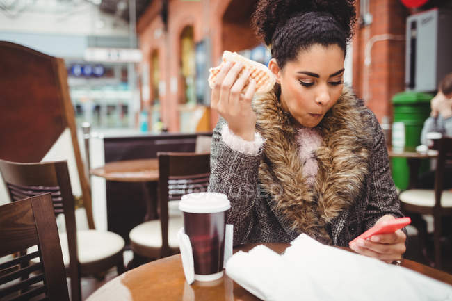 Mujer comiendo pan mientras usa el teléfono en el restaurante - foto de stock