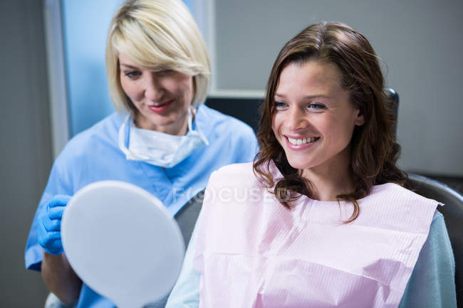 Lächelnde Patientin, die in den Spiegel schaut, während ein Zahnarzt im Bett sitzt — Stockfoto