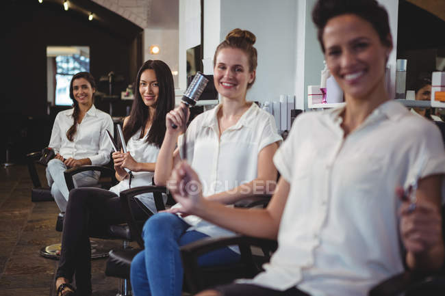 Retrato de sorrindo cabeleireiros multiculturais sentados em cadeiras no salão — Fotografia de Stock
