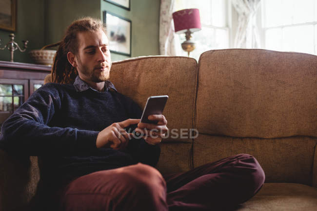 Joven hombre hipster utilizando el teléfono móvil en el sofá en casa - foto de stock