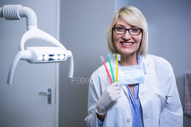 Портрет улыбающегося стоматолога с тремя зубными щетками в стоматологической клинике — стоковое фото